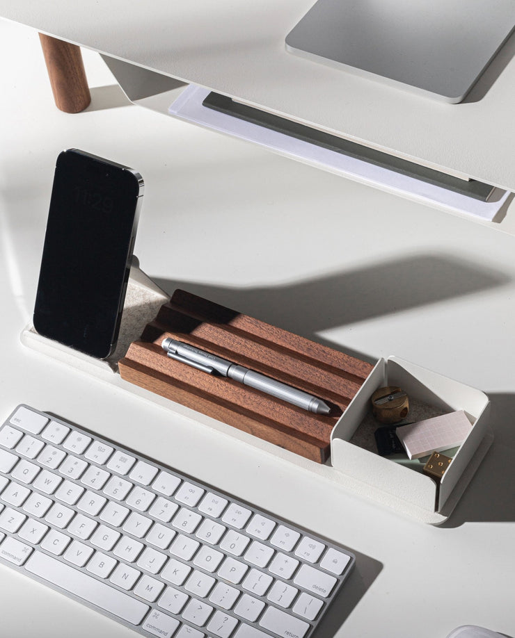 A minimalist white desk setup includes a wireless keyboard, silver pen on a walnut pen holder, smartphone, and the Gather Wood Zigzag Tray (2x1 - Walnut) holding keys and USB drives—all neatly arranged under bright lighting.