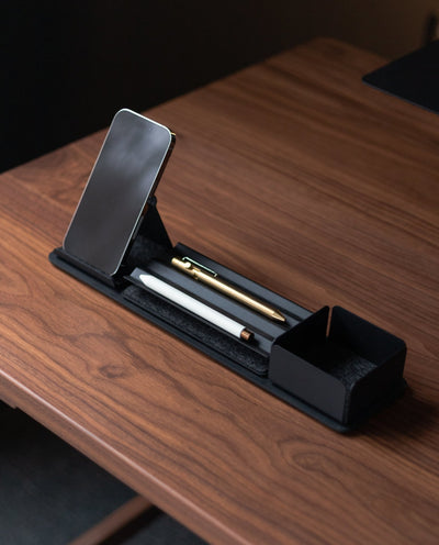 A wooden desk displays the Gather 1x4 Organizer Set (Black), holding a smartphone, white pen, gold pen, and an open compartment for storage, all in a sleek minimalist and modern style.