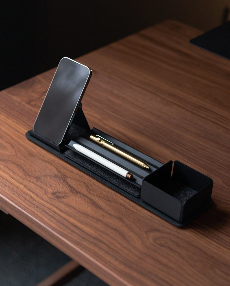 A wooden desk displays the Gather 1x4 Organizer Set (Black), holding a smartphone, white pen, gold pen, and an open compartment for storage, all in a sleek minimalist and modern style.