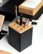 A black and wooden Warehouse Item - Gather Pencil Cup Lid (Maple), expertly crafted from solid maple wood in Pennsylvania, holds two pens, a pencil, a stylus, and an eraser. It is placed on a desk alongside a notepad and other office items in the background.