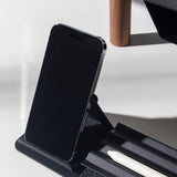 A black smartphone sits upright on the Gather Phone Stand (1x1 - Black) while a white stylus rests in its groove. Sunlight casts clear shadows across the scene.