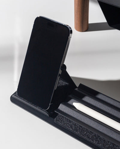 A black smartphone sits upright on the Gather Phone Stand (1x1 - Black) while a white stylus rests in its groove. Sunlight casts clear shadows across the scene.