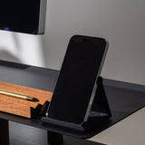 A smartphone in a black case rests on the Gather Phone Stand (1x1 - Black) on a desk beside a wooden pen holder with a gold pen.