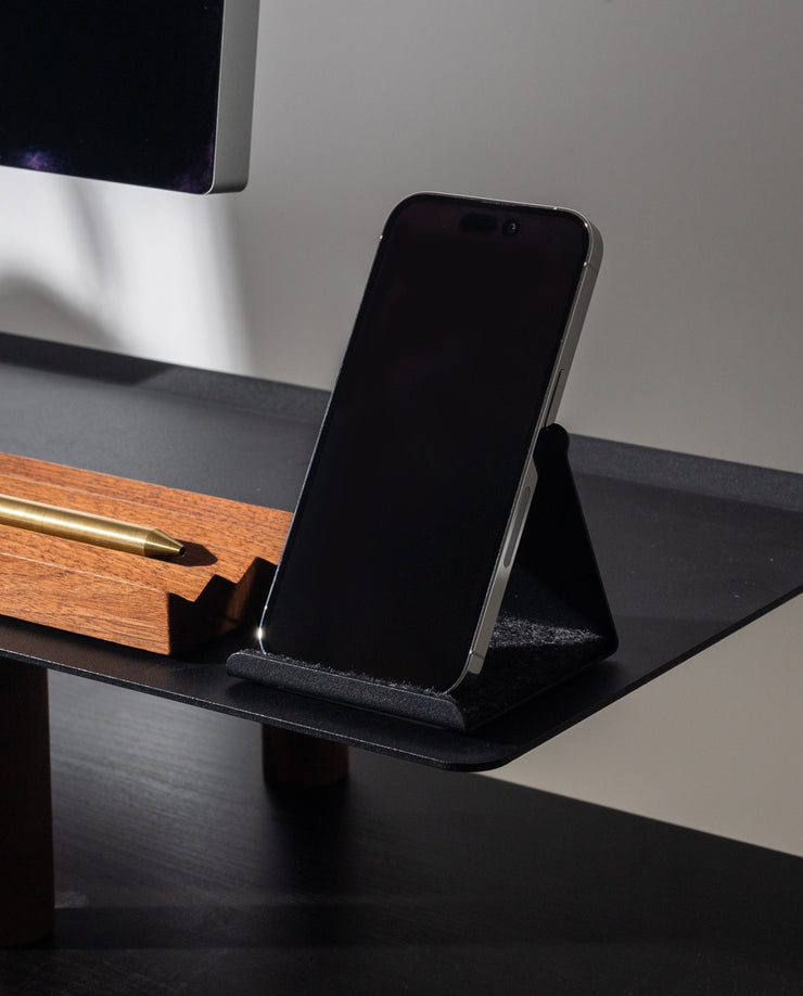 A smartphone in a black case rests on the Gather Phone Stand (1x1 - Black) on a desk beside a wooden pen holder with a gold pen.