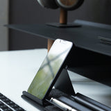 A Gather Phone Stand (1x1 - Black) holds a smartphone beside a keyboard and two pens on a white desk, with a black monitor stand and headphones in the background.