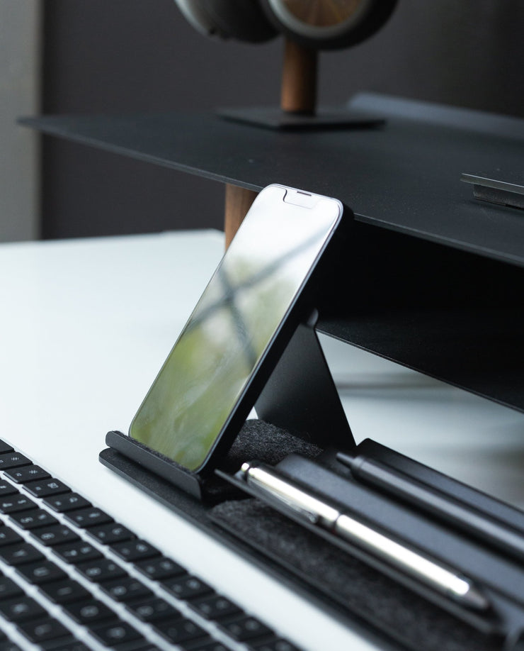 A Gather Phone Stand (1x1 - Black) holds a smartphone beside a keyboard and two pens on a white desk, with a black monitor stand and headphones in the background.