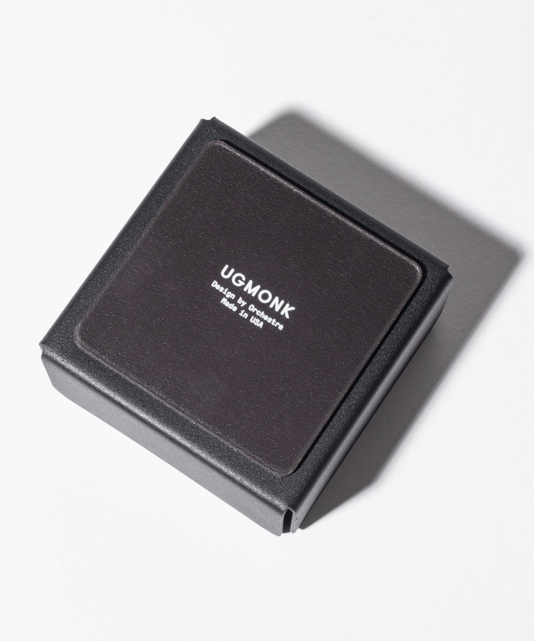 A Gather Small Tray (1x1 - Black), perfect as a desk organizer, featuring "UGMONK" and "Designed by Jeff Sheldon Made in USA" in white text on the lid, rests on a white surface with its shadow to the right. Proudly made in Pennsylvania.