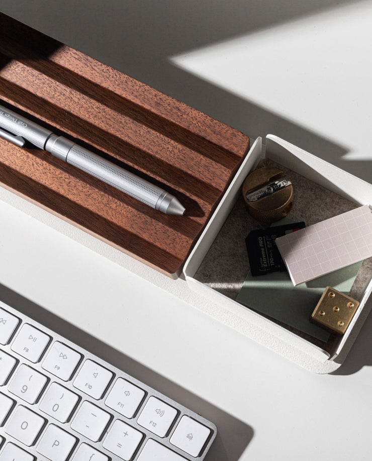 A close-up of a white desk with a keyboard, Gather Small Tray (1x1 - White) as a desk organizer holding a sharpener, eraser, SD card, paper clips, notepads, and wooden pen holder. Sunlight casts shadows across the items. Pen holder is made in Pennsylvania.