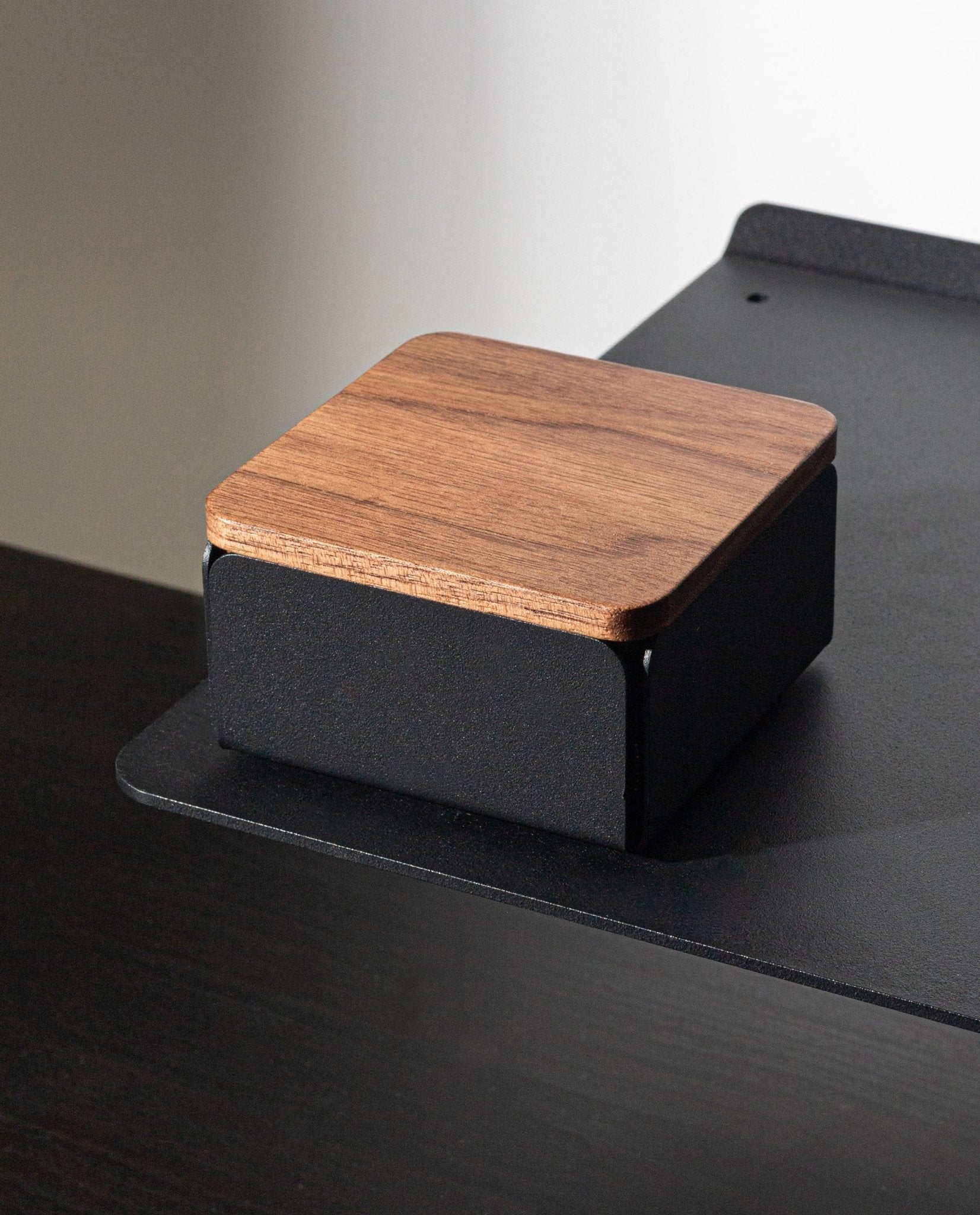A Gather Solid Wood Lid (Walnut) rests atop a small, square device with a black base, positioned on the edge of a black desk or shelf against a plain background.