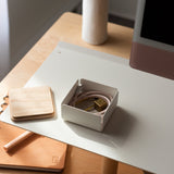 A tidy desk with a white monitor stand, Gather Small Tray (1x1 - White) desk organizer, light wooden coaster, closed tan notebook, and a small box with pink charging cable and gold plug, all lit by sunlight. Made in Pennsylvania.