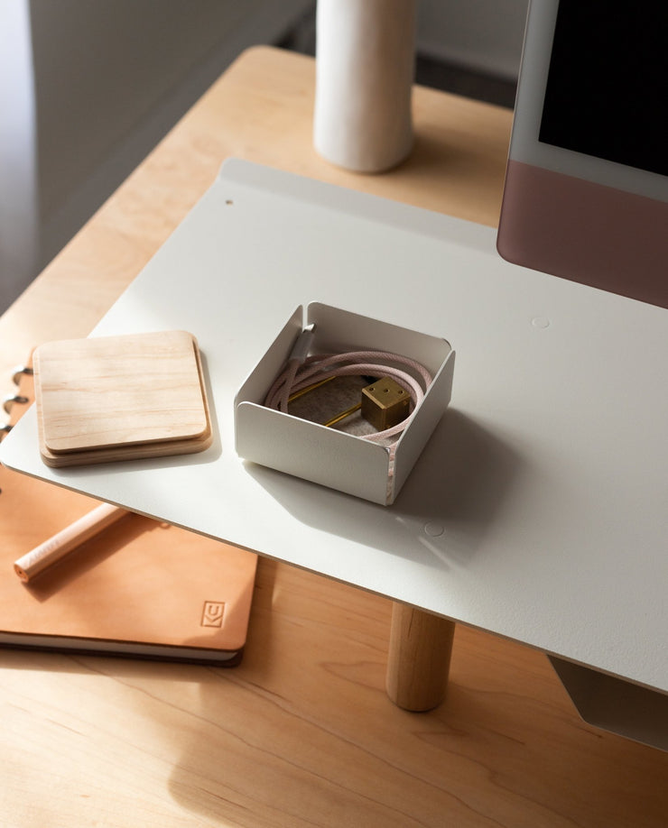 A tidy desk with a white monitor stand, Gather Small Tray (1x1 - White) desk organizer, light wooden coaster, closed tan notebook, and a small box with pink charging cable and gold plug, all lit by sunlight. Made in Pennsylvania.