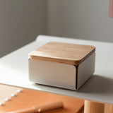 The Gather Small Tray (1x1 - White), a compact square organizer with a wooden lid, rests on a white surface. Crafted in Pennsylvania, its smooth lines and wood grain are softly lit. A tan cutting board and rolling pin appear in the background.