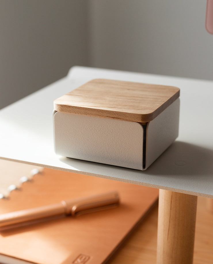The Gather Small Tray (1x1 - White), a compact square organizer with a wooden lid, rests on a white surface. Crafted in Pennsylvania, its smooth lines and wood grain are softly lit. A tan cutting board and rolling pin appear in the background.