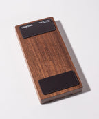 The Gather Wood Zigzag Tray (2x1 - Walnut) is a rectangular walnut block with rounded edges, "UGMONK" engraved in the center, and two black pads on each end, perfect for holding pens and pencils on your desk.