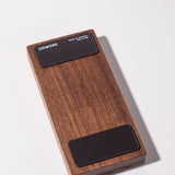 The Gather Wood Zigzag Tray (2x1 - Walnut) is a rectangular walnut block with rounded edges, "UGMONK" engraved in the center, and two black pads on each end, perfect for holding pens and pencils on your desk.