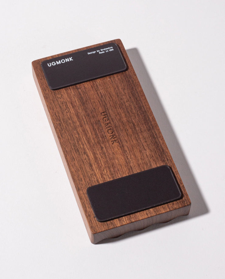 The Gather Wood Zigzag Tray (2x1 - Walnut) is a rectangular walnut block with rounded edges, "UGMONK" engraved in the center, and two black pads on each end, perfect for holding pens and pencils on your desk.