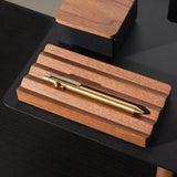 A brass pen sits on the Gather Wood Zigzag Tray (2x1 - Walnut), set on a black surface, with part of a matching pen and pencil holder in the background.