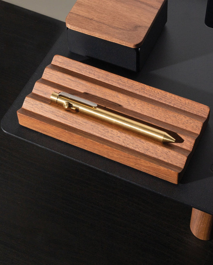 A brass pen sits on the Gather Wood Zigzag Tray (2x1 - Walnut), set on a black surface, with part of a matching pen and pencil holder in the background.
