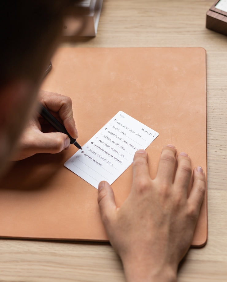 A person writes on an Analog - Task Cards (12-Pack), part of a productivity system, placed on a tan leather desk pad, with tasks like READ BOOK and BREAKFAST partially visible on the card.