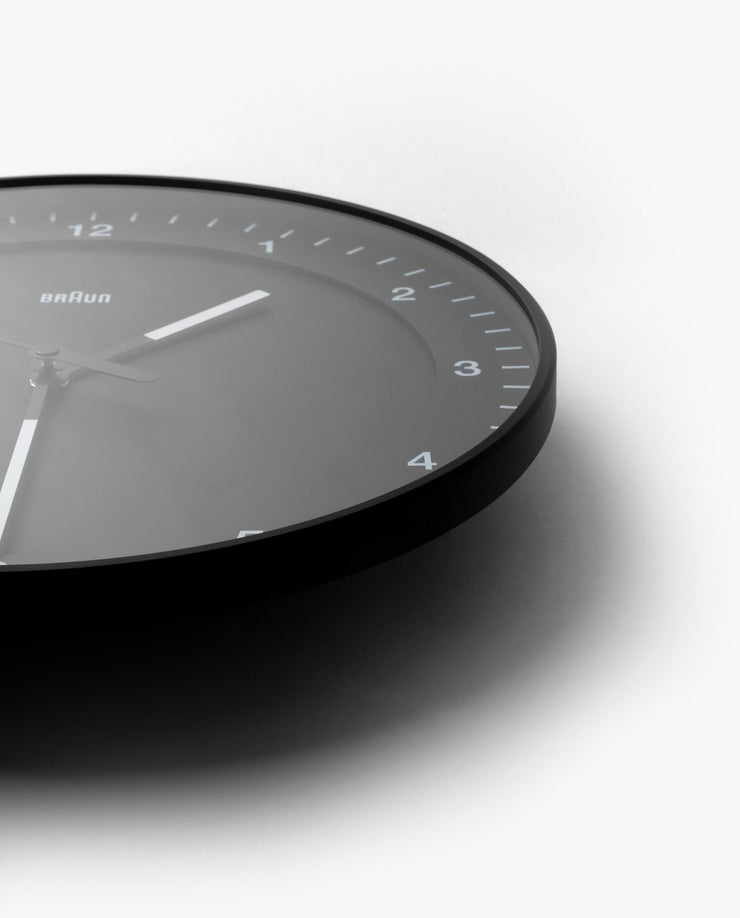 The Braun Wall Clock (Black) features minimalist white hour markers and hands, shown at an angle with only the lower right of the face visible against a white background. An easy-to-read clock, ideal for any modern space.