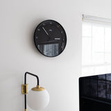 A modern, minimal office features the Braun Wall Clock (Black) displaying 12:14, a round wall lamp, part of a computer monitor, and a window letting in daylight.