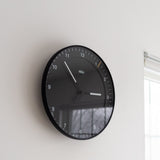 A Braun Wall Clock (Black) with white numbers and hands displays 10:10. Mounted on a white wall near a window, it’s easy to read in daylight streaming through.