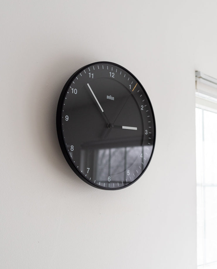 A Braun Wall Clock (Black) with white numbers and hands displays 10:10. Mounted on a white wall near a window, it’s easy to read in daylight streaming through.
