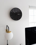 A Braun Wall Clock (Black) showing 10:08 hangs on a white wall above a globe-shaped lamp by a window. Its easy-to-read face and silent mechanism promote calm, with the edge of a computer monitor visible in the foreground.