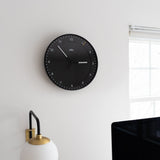 A Braun Wall Clock (Black) showing 10:08 hangs on a white wall above a globe-shaped lamp by a window. Its easy-to-read face and silent mechanism promote calm, with the edge of a computer monitor visible in the foreground.