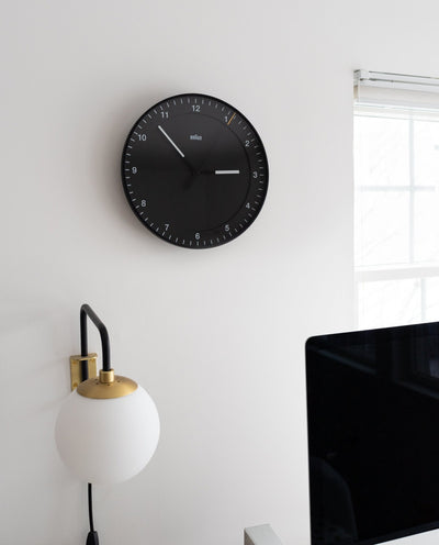 A Braun Wall Clock (Black) showing 10:08 hangs on a white wall above a globe-shaped lamp by a window. Its easy-to-read face and silent mechanism promote calm, with the edge of a computer monitor visible in the foreground.