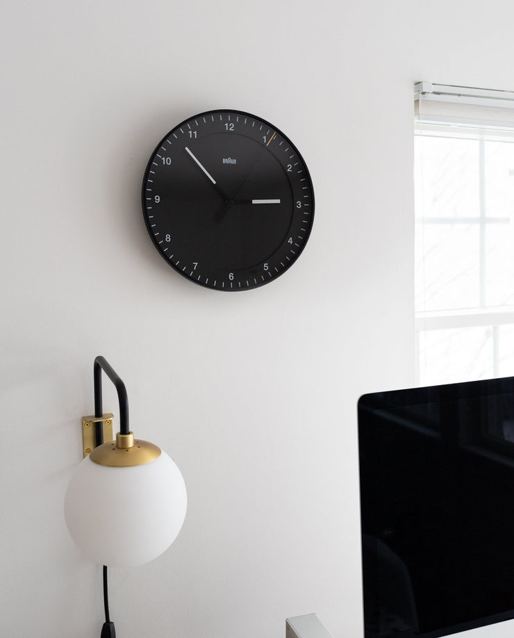 A Braun Wall Clock (Black) showing 10:08 hangs on a white wall above a globe-shaped lamp by a window. Its easy-to-read face and silent mechanism promote calm, with the edge of a computer monitor visible in the foreground.