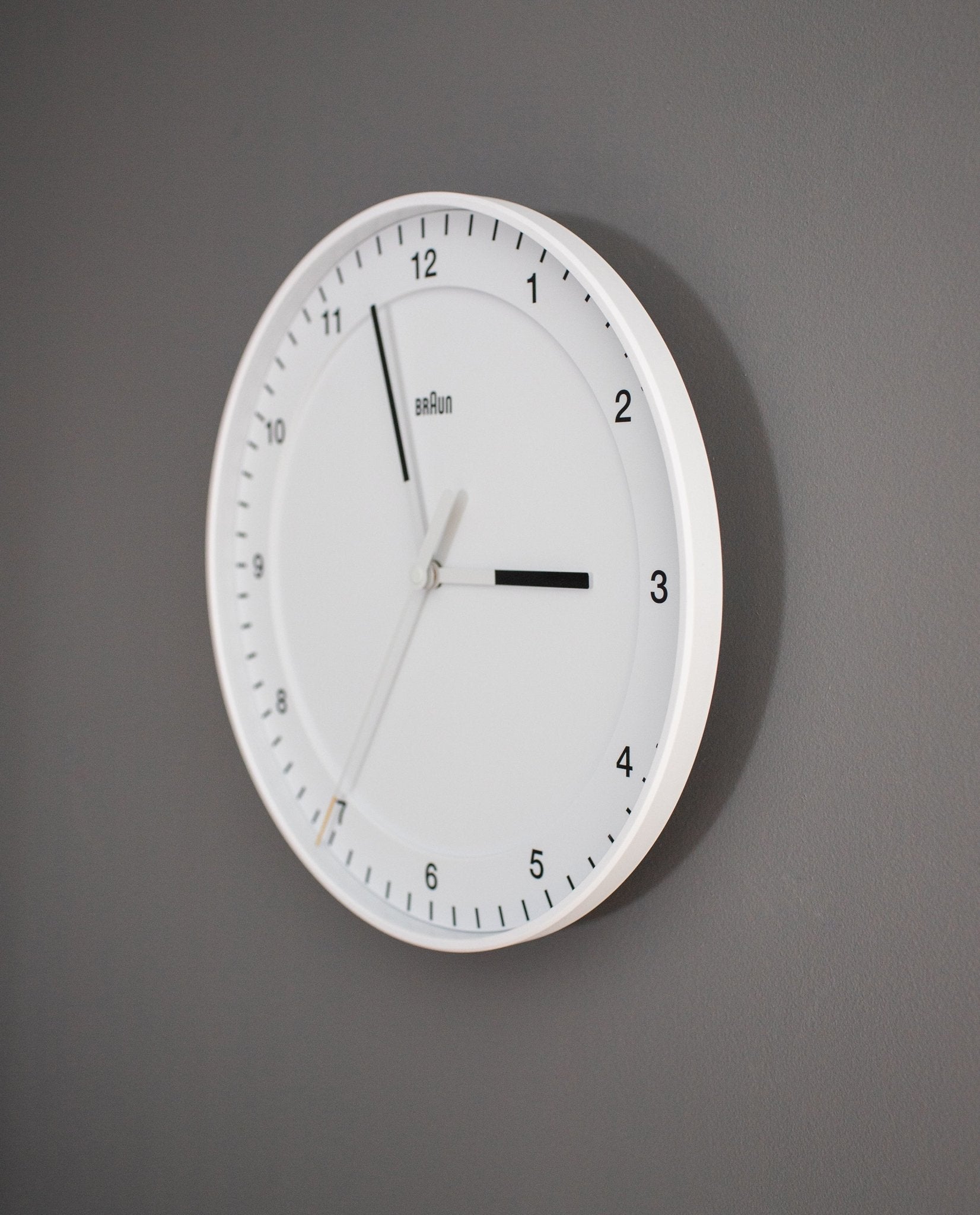 A Braun Wall Clock (White) with black hour and minute markers hangs on a gray wall, displaying the time 12:20:32. This clock showcases a minimalist design featuring thin black hands, a silent sweeping second hand, and an easy-to-read numberless dial.