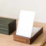 A wooden card holder displays a blank card from the Analog - Task Cards (12-Pack) set on a light wood desk. Stacked dark green boxes and a gold pen are nearby. Text below notes: Analog Wood Card Holder sold separately.