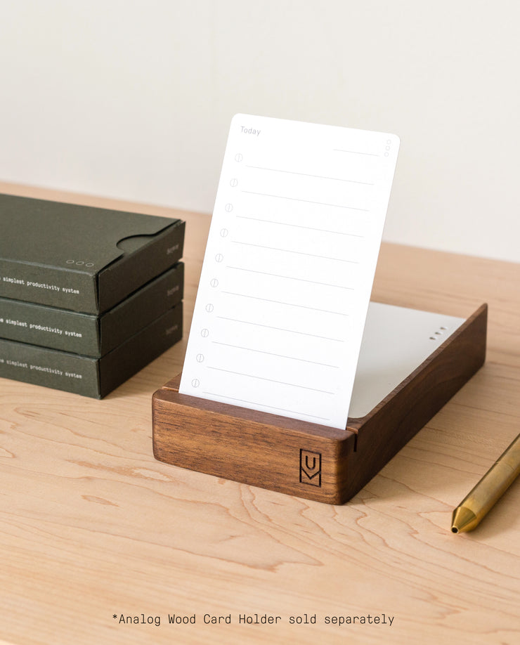 A wooden card holder displays a blank card from the Analog - Task Cards (12-Pack) set on a light wood desk. Stacked dark green boxes and a gold pen are nearby. Text below notes: Analog Wood Card Holder sold separately.
