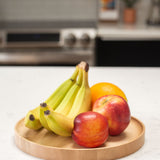 A Round Valet Tray (Maple - Large), handcrafted in Pennsylvania, sits on a white countertop holding three bananas, an orange, a red apple, and a nectarine. A blurred kitchen stove and other items are visible in the background.