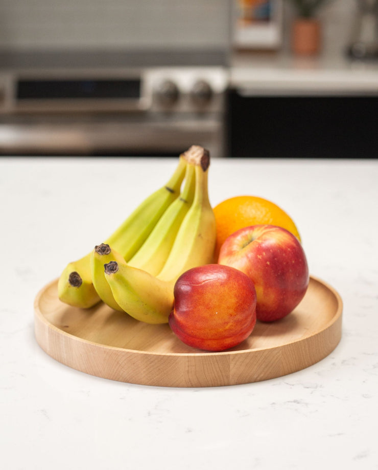 A Round Valet Tray (Maple - Large), handcrafted in Pennsylvania, sits on a white countertop holding three bananas, an orange, a red apple, and a nectarine. A blurred kitchen stove and other items are visible in the background.