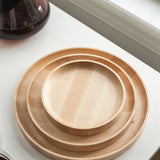 A handcrafted Round Valet Tray (Maple - Large), made in Pennsylvania, is stacked with two smaller maple trays on a white surface near a window, alongside part of a glass pitcher and a cup.