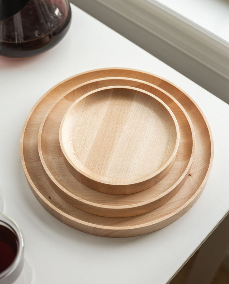 A handcrafted Round Valet Tray (Maple - Large), made in Pennsylvania, is stacked with two smaller maple trays on a white surface near a window, alongside part of a glass pitcher and a cup.