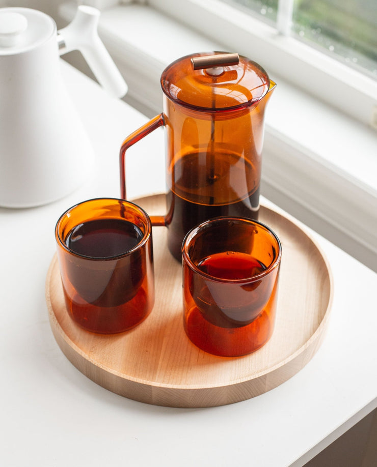 A handcrafted Round Valet Tray (Maple - Large), made in Pennsylvania, holds a transparent amber glass French press and two matching cups filled with dark coffee, placed on a white surface near a window.