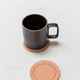 A Hasami Porcelain Mug (Matte Black) sits on a round tan coaster, with another nearby embossed with "ENJOY THE JOURNEY," all resting on a white marble surface.