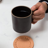 A hand holding a Hasami Porcelain Mug (Matte Black) above a round tan coaster embossed with "ENJOY THE JOURNEY," set on a white surface.