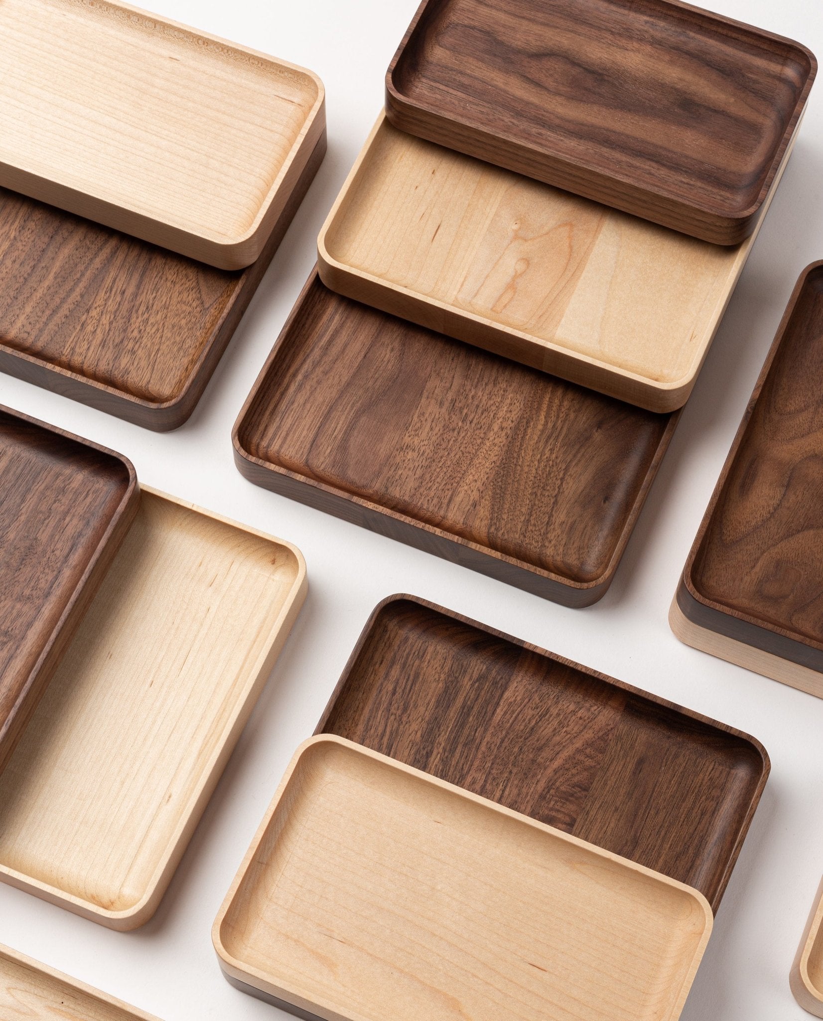 Several rectangular wooden trays, including the handcrafted Valet Tray (Maple - Small) in solid maple, as well as trays in other light and dark wood tones, are arranged in an overlapping, grid-like pattern on a white surface.