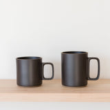 Two Hasami Porcelain Mugs (Matte Black) with square handles sit side by side on a light wooden shelf against a plain white background, one slightly shorter and wider than the other, showcasing subtle handmade charm.