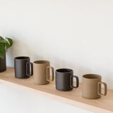 Four Hasami Porcelain Mugs (Matte Black), inspired by handmade Japanese design, alternate with beige mugs on a light wooden shelf against a white wall. A black-potted plant partially appears on the shelf’s left side.