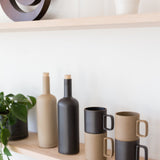 Two Hasami Porcelain Mugs (Matte Black) are stacked on a light wooden shelf beside two bottles with cork stoppers and a leafy green potted plant, all set against a white wall.