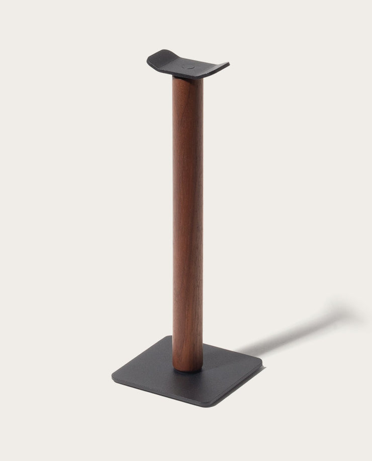 The Gather Headphone Stand (1x1 - Black/Walnut) features a solid walnut vertical column, a black powder coated steel base, and a curved top to hold headphones, all set against a plain light background.