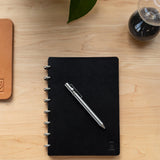 A black spiral-bound notebook with the INVENTERY Bolt Action Pen (Stainless) placed diagonally on top sits on a light wooden desk. A portion of a tan leather item, a plant leaf, and a glass hourglass are also visible.