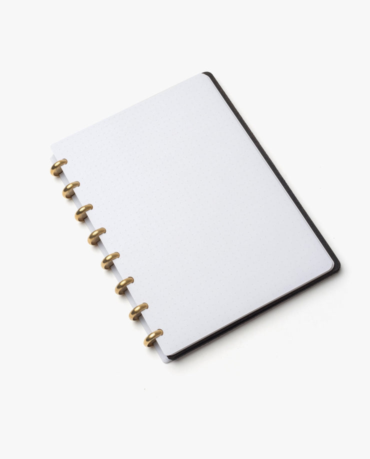 A Discbound Heirloom Journal Bundle (Black Leather - Standard 6" x 8.5" + 3 Refills) with refillable white dotted pages and gold metal rings rests on a white surface, viewed from a slight angle.