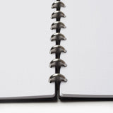 A close-up, low-angle view of the Discbound Heirloom Journal Bundle (Black Leather - Standard 6" x 8.5" + 3 Refills), featuring dotted pages and central metal disc binding connecting both sides of this sleek refillable notebook.