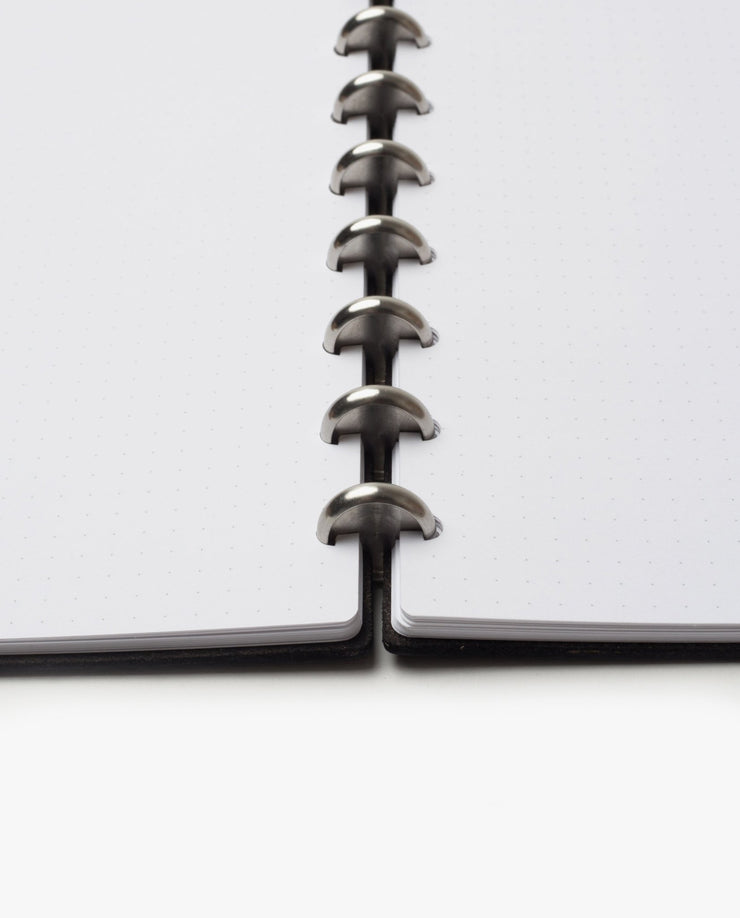 A close-up, low-angle view of the Discbound Heirloom Journal Bundle (Black Leather - Standard 6" x 8.5" + 3 Refills), featuring dotted pages and central metal disc binding connecting both sides of this sleek refillable notebook.